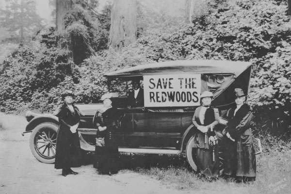 An old car with save the redwoods on it and women standing around it in dresses